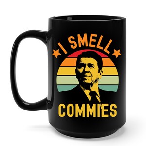 May include: Black ceramic mug with a handle. The mug features a graphic of Ronald Reagan in front of a retro sunset design. The text "I SMELL COMMIES" is printed in orange and yellow.