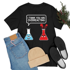 Puede incluir: Camiseta negra con un gráfico de temática científica. El gráfico presenta dos vasos de dibujos animados, uno azul y sonriente, el otro rojo y enfadado, con el texto "I THINK YOU ARE OVERREACTING". También se muestran unos vaqueros azules, un gorro beige y zapatos negros.