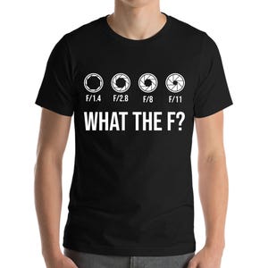 Könnte beinhalten: Schwarzes T-Shirt mit einer weißen Grafik von vier Kamera-Blendenstufen, beschriftet mit F/1.4, F/2.8, F/8 und F/11. Der Text "WHAT THE F?" befindet sich unterhalb der Blendenstufen.