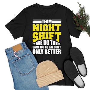 Puede incluir: Camiseta negra con el texto "TEAM NIGHT SHIFT WE DO THE SAME JOB AS DAY SHIFT ONLY BETTER" en amarillo y blanco. La camiseta se combina con vaqueros azules, un gorro beige y zapatos negros.