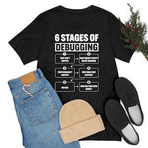May include: Black t-shirt with the text "6 Stages of Debugging" and a flowchart of debugging stages. The shirt is paired with blue jeans, a beige beanie, and black shoes. A candy cane is in the jeans pocket.