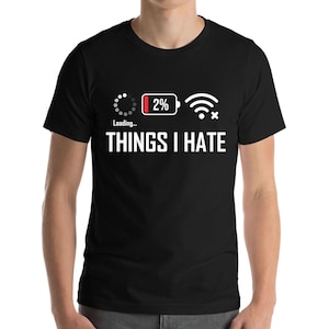 Peut inclure: T-shirt noir avec l'inscription blanche « THINGS I HATE ». Au-dessus du texte, des graphiques d'un symbole de chargement, d'une batterie à 2 % et d'un symbole Wi-Fi barré. Le t-shirt est à manches courtes.
