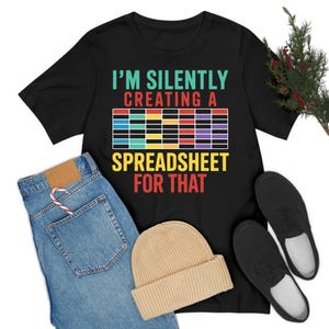 Puede incluir: Camiseta negra con el texto "I'M SILENTLY CREATING A SPREADSHEET FOR THAT" en turquesa, rojo, amarillo y morado. La camiseta se combina con vaqueros azules, un gorro beige y zapatos negros.