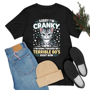 May include: Black t-shirt with a graphic of a grumpy gray tabby cat and the text "SORRY I'M CRANKY I'M GOING THROUGH MY TERRIBLE 60'S RIGHT NOW". The image also includes blue jeans, a tan beanie, and black shoes.