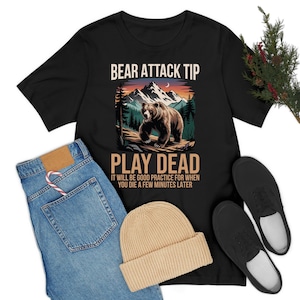 Puede incluir: Camiseta negra con un gráfico de un oso en un entorno de bosque. El texto "BEAR ATTACK TIP PLAY DEAD IT WILL BE GOOD PRACTICE FOR WHEN YOU DIE A FEW MINUTES LATER" está impreso en la camiseta.