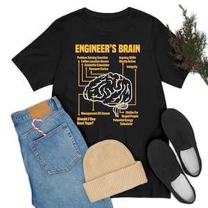 Op de afbeelding: Zwart T-shirt met een gele afbeelding van een brein met het opschrift "Engineer's Brain" met humoristische engineering-gerelateerde functies. De afbeelding bevat een blauwe jeans, een beige muts en zwarte schoenen.