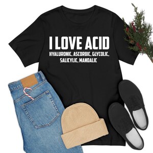 Op de afbeelding: Zwart T-shirt met de tekst "I LOVE ACID" en de namen van verschillende zuren. Het shirt wordt gecombineerd met een blauwe jeans, een beige muts en zwarte sneakers.
