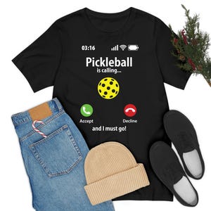 Może przedstawiać: Czarny T-shirt z białą grafiką telefonu. Ekran telefonu wyświetla napis "Pickleball is calling..." z żółtą ikoną pickleballa. Telefon ma zielone przyciski "Zaakceptuj" i czerwone "Odrzuć". Tekst pod telefonem brzmi "and I must go!"