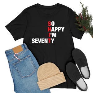 Könnte beinhalten: Schwarzes T-Shirt mit dem Text "SO HAPPY I'M SEVENTY" in weißer und roter Schrift. Das Shirt wird mit einer blauen Jeans, einer beigen Mütze und schwarzen Schuhen kombiniert. Eine Zuckerstange befindet sich in der Jeans-Tasche.