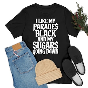 Peut inclure: T-shirt noir avec l'inscription blanche "I LIKE MY PARADES BLACK AND MY SUGARS GOING DOWN". Le t-shirt est associé à un jean bleu, un bonnet beige et des chaussures noires. Un sucre d'orge est dans la poche du jean.
