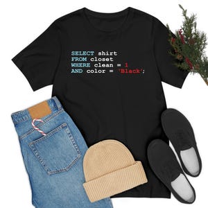 May include: Black t-shirt with the text "SELECT shirt FROM closet WHERE clean = 1 AND color = 'Black';". The image also shows blue jeans, a tan beanie, and black shoes.