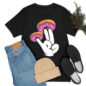 May include: Black t-shirt with a white hand holding two pink and yellow donuts. The hand is making a peace sign.