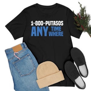 May include: Black t-shirt with the text "1-800-PUTASOS ANY TIME WHERE" in white and blue. The shirt is paired with blue jeans, a tan beanie, and black shoes. A candy cane is in the jeans pocket.