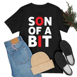 May include: Black t-shirt with the text "SON OF A BIT" in white and red. The image also includes a pair of blue jeans, a tan beanie, and black shoes.