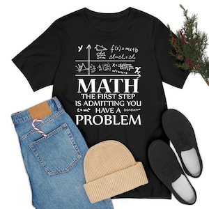 May include: Black t-shirt with white text and mathematical equations. The text reads "MATH THE FIRST STEP IS ADMITTING YOU HAVE A PROBLEM." Also shown are blue jeans, a beige beanie, and black shoes.