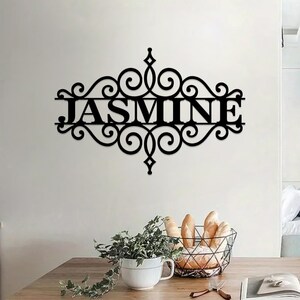 Pu&ograve; includere: Decorazione da parete in metallo nero con un design a volute decorative e il nome "JASMINE" al centro.