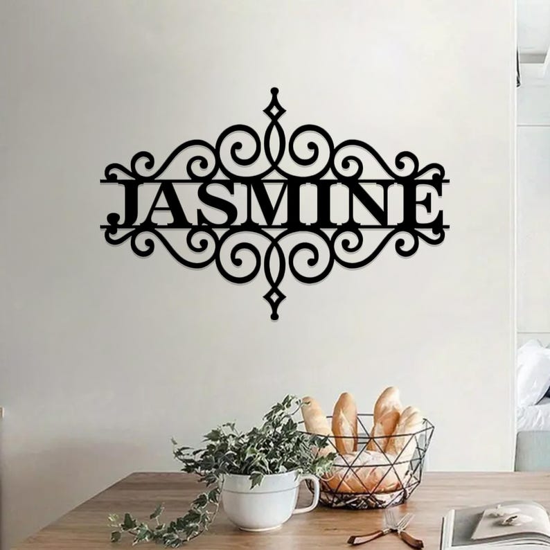 Pu&ograve; includere: Decorazione da parete in metallo nero con un design a volute decorative e il nome "JASMINE" al centro.