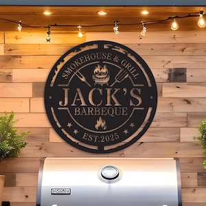 May include: A black metal sign with the words "Smokehouse & Grill" and "Jack's Barbeque" in a circular design. The sign includes a grill graphic, stars, and the year "Est. 2025". The sign is mounted on a wooden wall above a stainless steel grill.