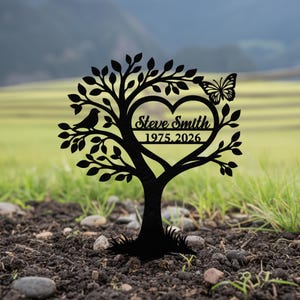 Puede incluir: Silueta de árbol de metal negro con un recorte en forma de corazón, un pájaro y una mariposa. El corazón contiene el nombre "Steve Smith" y los años "1975.2026". El árbol está plantado en tierra con hierba al fondo.