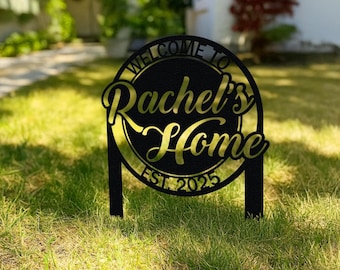 Puede incluir: Letrero de jardín de metal negro con el texto "WELCOME TO Rachel's Home EST. 2025" en letras doradas. El letrero es circular con un borde decorativo y está colocado en un jardín cubierto de hierba.