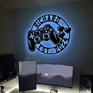 Könnte beinhalten: Eine schwarze und weiße Wanddekoration aus Metall mit einem Videospiel-Controller und dem Text "Richard Est. 2022" darum herum. Die Dekoration wird mit blauem Licht beleuchtet.
