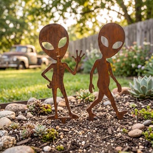May include: Two rust-colored metal alien silhouettes standing in a garden. One alien figure is waving with its left hand, while the other is depicted in a walking pose. Both figures feature large, oval-shaped eye cutouts and slender limbs.