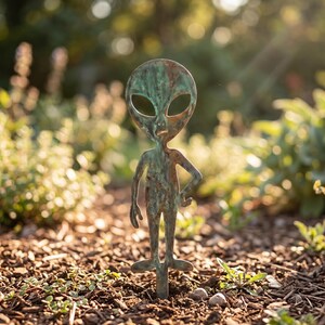 Könnte beinhalten: Ein Gartenstecker aus Metall in Form einer Alien-Figur mit einer verwitterten, oxidierten grün-braunen Patina. Die Figur hat einen großen, ovalen Kopf mit hohlen Augenhöhlen, einen schlanken Oberkörper und eine Hand in der Hüfte.