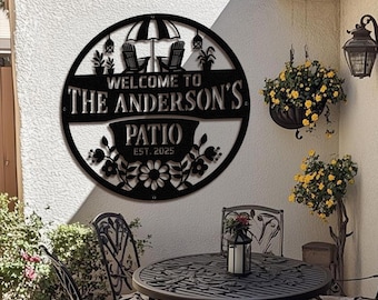 May include: Black metal patio sign with the words "Welcome to The Anderson's Patio Est. 2025". The sign features an umbrella, two chairs, and floral accents. The sign is round and mounted on a white wall.