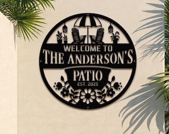 May include: Black metal patio sign with the text "WELCOME TO THE ANDERSON'S PATIO EST. 2025". The sign features a circular design with an umbrella, two chairs, plants, and floral accents. It is mounted on a light-colored wall.