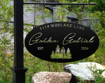 May include: Black metal oval sign with the text "1114 Woodland Lane Garden Gabriel EST 2024" and a silhouette of pine trees. The sign is hanging from a black metal rod attached to a black post.