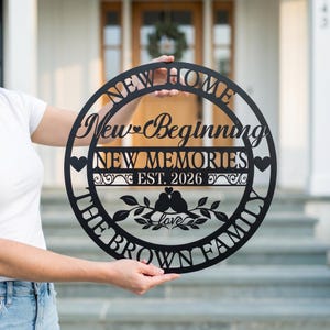 May include: Black metal sign with the words "NEW HOME, New Beginning, NEW MEMORIES, EST. 2026, THE BROWN FAMILY." The sign has a circular shape with decorative elements, including hearts and birds on a branch. The sign is held by a person.