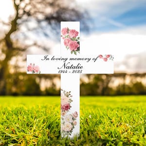 May include: White wooden cross with floral detailing and the inscription "In loving memory of Natalie 1944-2025." Pink and cream roses decorate the cross, set against a backdrop of green grass and a blurred sky.