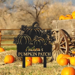 May include: A black metal sign with a pumpkin design and the text "Anthea's PUMPKIN PATCH" in gold lettering. The sign is in a field with pumpkins and a wooden wagon wheel.