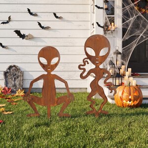 Könnte beinhalten: Zwei rostfarbene Alien-Silhouetten aus Metall auf einem grünen Rasen. Ein Alien hockt mit den Händen auf den Knien, während das andere aufrecht mit gewellten Armen steht. Beide Figuren haben große, oval geformte Aussparungen für die Augen.