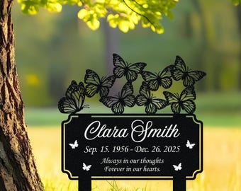 May include: Black metal memorial garden stake with butterfly accents. The sign reads "Clara Smith" with birth and passing dates, and the phrases "Always in our thoughts" and "Forever in our hearts."