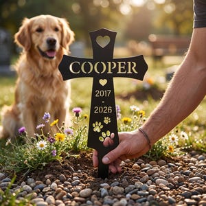 Puede incluir: Una estaca conmemorativa negra en forma de cruz para mascotas con el nombre "COOPER", las fechas 2017-2026 y huellas de patas. Un perro golden retriever está en el fondo. La estaca está colocada en un jardín con flores.