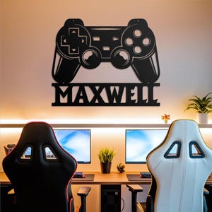May include: Black metal wall art in the shape of a video game controller with the name "MAXWELL" below it. The controller is mounted on a wall above two gaming chairs and computer monitors.