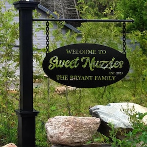 Può includere: Cartello in metallo nero con la scritta "WELCOME TO Sweet Nuzzles" e "THE BRYANT FAMILY EST. 2025". Il cartello ovale è appeso a un palo nero con catene. Lo sfondo presenta fogliame verde e rocce.