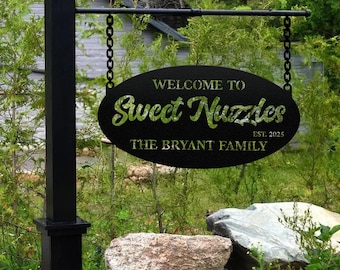 May include: Black metal sign with the text "WELCOME TO Sweet Nuzzles" and "THE BRYANT FAMILY EST. 2025". The sign is oval-shaped and hangs from a black post with chains. The background features green foliage and rocks.