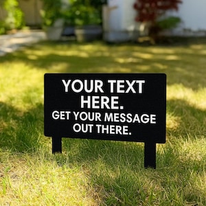 Könnte beinhalten: Schwarzes rechteckiges Gartenschild mit weißem Text: "YOUR TEXT HERE. GET YOUR MESSAGE OUT THERE." Das Schild steht im Gras.