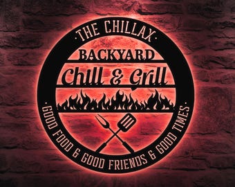 Könnte beinhalten: Schwarzes Metallschild mit dem Text "The Chillax Backyard Chill & Grill. Good Food & Good Friends & Good Times." Das Schild hat einen roten Schein dahinter und zeigt eine Silhouette von Flammen und gekreuzten Grillwerkzeugen.