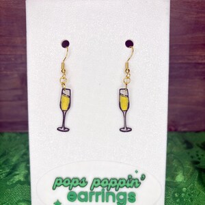 May include: A pair of gold-colored dangle earrings featuring a champagne glass design. The glasses are filled with yellow liquid and outlined in black. The earrings are displayed on a white card with the text "pops poppin' earrings".