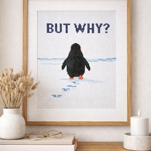 Op de afbeelding: In een houten lijst geplaatst kruissteekkunstwerk met een zwarte pinguïn op een witte achtergrond met de marineblauwe tekst "BUT WHY?". De pinguïn heeft oranje voeten en loopt op een besneeuwd oppervlak, waarbij hij voetstappen achterlaat.