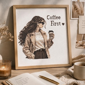 Puede incluir: Una obra de arte en punto de cruz enmarcada representa a una mujer con gafas de sol sosteniendo una taza de café, con el texto "Coffee First". La obra de arte está en un marco de madera, con una paleta de colores neutros. La imagen está sobre una superficie de madera.