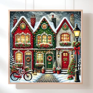 Puede incluir: Una obra de arte enmarcada con temática navideña representa tres casas cubiertas de nieve en rojo, verde y blanco, adornadas con coronas y luces. Una bicicleta roja con una cesta está frente a la primera casa. También se ve una farola y un árbol de Navidad.
