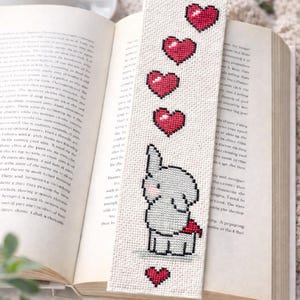 Peut inclure: Marque-page fait main avec un motif au point de croix. Il présente un éléphant gris tenant un cœur rouge, avec cinq cœurs rouges au-dessus. Le marque-page est inséré dans un livre ouvert, un accessoire charmant.