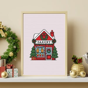 May include: Framed cross-stitch art showing a red Christmas bakery with a snow-covered roof. The word "BAKERY" is above the door. Two gingerbread men are in the window, with Christmas trees on either side. A wreath and gifts are also visible.