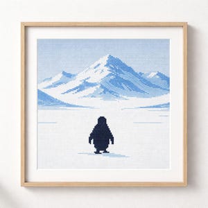 Può includere: Opera d'arte a punto croce di un pinguino in piedi su un paesaggio innevato con montagne sullo sfondo. L'opera è incorniciata in una cornice di legno chiaro. La tavolozza dei colori è principalmente blu e bianco.