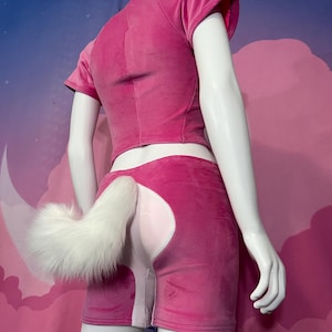 以下が含まれることがあります： マネキンのピンク色のベロアの衣装。半袖のクロップトップとマッチするショートパンツで構成されています。ふわふわの白い尻尾がショートパンツに付いています。背景はピンクと青の壁画です。