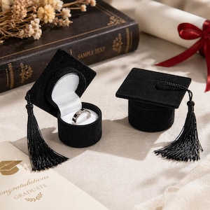 May include: Black velvet graduation cap ring boxes, one open to reveal a silver ring. Each box has a black tassel. A graduation card and a rolled diploma with a red ribbon are in the background.